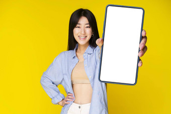 亚洲女孩持有智能手机显示白色<strong>屏</strong>幕兴奋微笑相机穿蓝色的衬衫孤立的黄色的<strong>背景</strong>伟<strong>大</strong>的提供移动应用程序广告产品放置