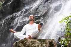 健康瑜伽冥想概念年轻的男人。坐着莲花位置岩石热带瀑布