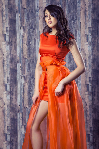 年轻的浅黑肤色的女人女人时尚的橙色丝绸衣服摆姿势木背景
