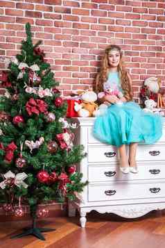 美丽的十几岁的金发女郎坐白色床头柜上圣诞节树玩具礼物