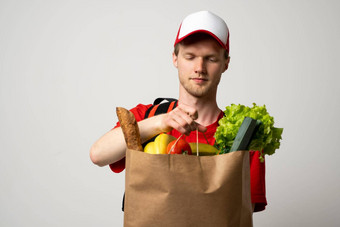 杂货店交付快递男人。红色的帽t恤热袋肩膀持有纸袋食物白色工作室背景食物交付服务在线购物