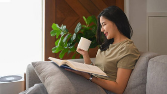 和平年轻的亚洲女人喝Herbal茶阅读书舒适的沙发上首页休闲活动概念