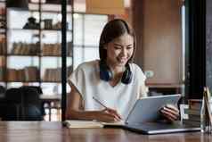 亚洲女学生数字平板电脑研究远程听在线教训采取笔记笔记本坐着咖啡商店在线教育