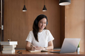 快乐亚洲女人坐桌子上首页写研究在线电脑微笑年轻的混合比赛女工作遥远的移动PC网络教育学习概念
