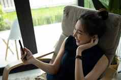 快乐的年轻的女人放松舒适的扶手椅使视频调用聪明的电话