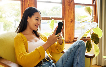 拉美裔女人智能手机坐着前面窗口明亮的生活房间年轻的快乐女微笑手机放松椅子现代技术首页封锁