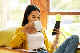 女人智能手机喝咖啡坐着前面窗口明亮的生活房间年轻的女手机放松椅子现代技术首页