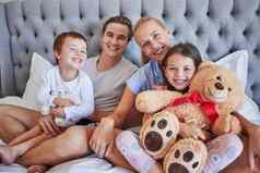 肖像快乐高加索人家庭孩子们坐着床上持有Teddybear微笑相机爱的父母支出免费的时间女儿儿子周末