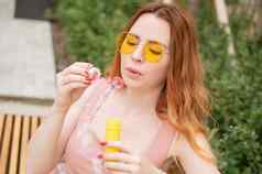 年轻的红发女人吹肥皂泡沫在户外女孩黄色的太阳镜牙套