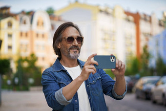 快乐中间岁的灰色有胡子的男人。采取图片智能手机相机小镇穿休闲快乐高级男人。街欧洲城市移动电话旅行概念