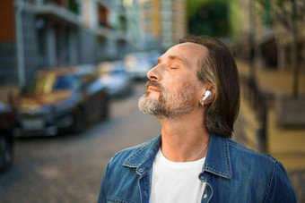 享受生活听音乐眼睛关闭站在户外小镇街英俊的成熟的灰色有胡子的男人。无线耳机穿休闲装旅行生活方式概念