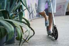 关闭男人。骑小镇成人踏板车大轮子时尚的运动鞋街道公园长剑长龙舌兰植物工作在户外脸可见