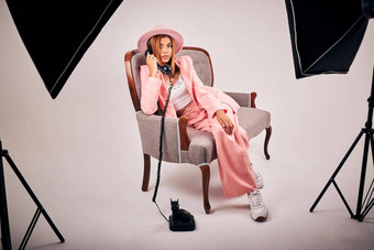 时尚的年轻的混合比赛女摆姿势时尚的粉红色的衣服坐着<strong>椅子</strong>使调用<strong>复古</strong>的电话工作室拍的女模型集编辑时尚拍摄