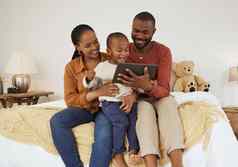 可爱的非洲美国男孩数字平板电脑坐着床上父母年轻的妈妈。父亲看兴奋蹒跚学步的儿子戏剧游戏看卡通电影在线