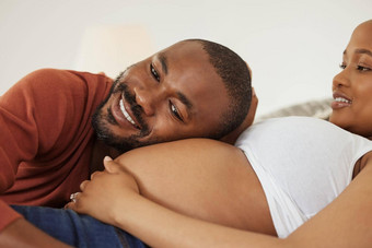快乐年轻的非洲美国父亲听怀孕了的妻子肚子微笑年轻的男人。把头准的妻子肚子听运动
