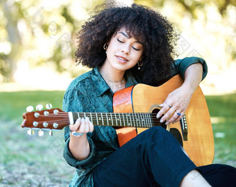 年轻的混合比赛女人卷曲的非洲式发型坐着公园玩吉他年轻的快乐拉美裔女人玩吉他坐着自然放松玩吉他
