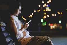 年轻的女人智能手机坐板凳上晚上模糊灯