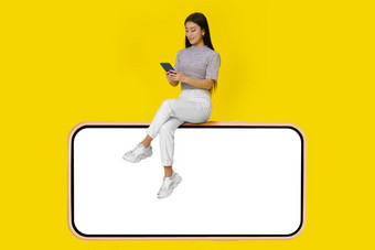 亚洲女孩电话手坐巨大的智能手机白色屏幕穿休闲白色牛仔裤孤立的黄色的背景模拟产品放置移动应用程序广告复制空间