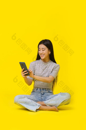 亚洲年轻的女孩地板上电话手发短信购物在线玩游戏孤立的黄色的背景移动应用程序广告产品放置复制空间