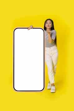 发送空气吻亚洲女孩站巨大的巨大的智能手机白色屏幕穿着休闲孤立的黄色的背景免费的空间模拟移动应用程序广告
