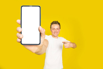 移动应用程序广告英俊的金发碧眼的男人。指出巨大的智能手机白色空屏幕穿白色t恤牛仔裤孤立的黄色的背景伟大的提供