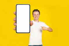 英俊的金发碧眼的男人。指出巨大的智能手机白色空屏幕穿白色t恤牛仔裤手机显示模拟孤立的黄色的背景移动应用程序广告