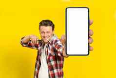 快乐英俊的男人。指出巨大的智能手机白色空屏幕穿红色的格子衬衫手机显示模拟孤立的黄色的背景移动应用程序广告