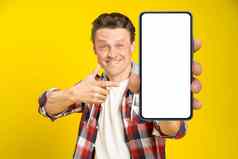 迷人的快乐男人。指出智能手机白色空屏幕穿红色的格子衬衫牛仔裤手机显示模拟孤立的黄色的背景移动应用程序广告