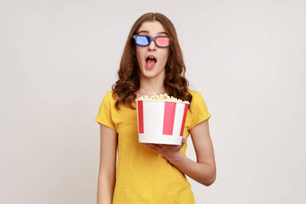 惊讶年轻的女imax眼镜看电影电影持有爆米花看令人惊异的电影保持口开放穿黄色的t恤