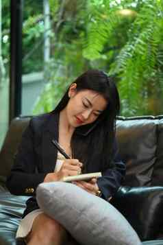 漂亮的年轻的女商人坐着沙发个人办公室写作业务计划笔记本
