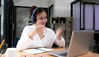 微笑年轻的亚洲女人耳机波问候会说话的网络摄像头虚拟谈话移动PC快乐女无线耳机说话视频调用电脑咨询客户端在线
