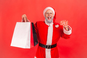 肖像快乐满意上了年纪的男人。灰色的胡子穿圣诞老人老人服装持有购物袋出售卡片购物中心折扣