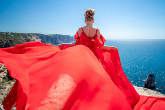 金发女郎长头发阳光明媚的海滨红色的流动衣服回来视图丝绸织物挥舞着风背景蓝色的天<strong>空山</strong>海滨