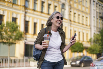 欧洲高级女人灰色的头发快乐享受免费的时间工作旅行持有电话汁塑料杯城市背景享受生活成熟的女人