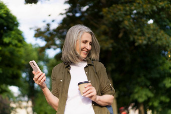 成熟的欧洲女人灰色的头发快乐享受免费的时间工作旅行持有电话<strong>饮料</strong>咖啡<strong>纸杯</strong>城市花园公园享受生活成熟的女人