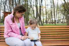 女孩运营智能手机坐着公园板凳上照顾者