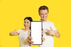持有智能手机白色屏幕高加索人男人。亚洲女人指出手指兴奋移动应用程序广告孤立的黄色的背景产品放置