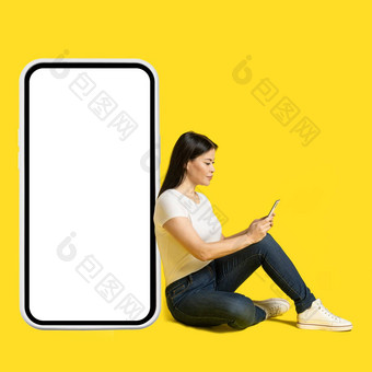 坐着地板上中间岁的亚洲女人电话手靠巨大的巨大的智能手机白色屏幕穿白色t恤牛仔裤孤立的黄色的背景免费的空间模拟