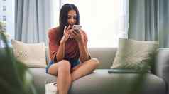 有吸引力的年轻的混合比赛女人坐着沙发上智能手机浏览互联网移动应用程序消息传递社会网络首页