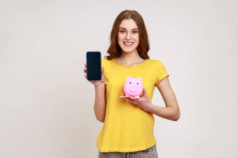 年轻的有吸引力的女人持有小猪银行智能手机空白屏幕促销活动微笑艺术相机穿黄色的t恤