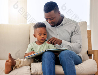 非洲美国爸爸指出图片故事书可爱的男孩坐着父亲阅读故事书沙发上首页父亲儿子支出时间