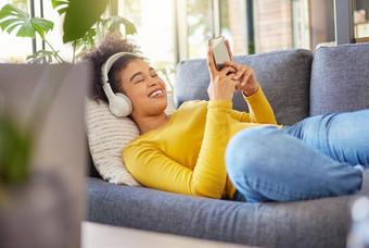 年轻的快乐的混合比赛女人穿耳机听音乐打字消息电话首页内容拉美裔女卷曲的非洲式发型社会媒体手机放松沙发上首页