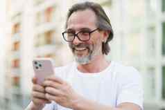 快乐高加索人中间岁的男人。灰色胡子阅读发短信智能手机站在户外城市街道背景穿白色衬衫中间岁的男人。街欧洲城市