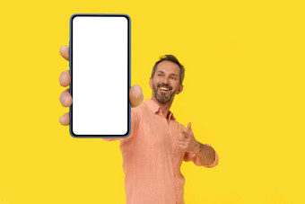 中间岁的灰色头发的男人。指出智能手机手惊讶微笑智能手机穿桃子衬衫孤立的黄色的成熟的适合男人。电话移动应用程序广告