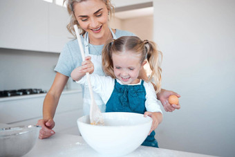 快乐妈妈。女儿烘焙成键年轻的女人帮助女儿烤首页微笑妈妈。持有蛋烹饪女儿快乐混合碗面糊