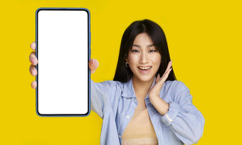 美丽的亚洲女孩凭借自己的面部表达式持有智能<strong>手机</strong>白色屏幕快乐介绍应用程序游戏赢得孤立的黄色的背景产品放置移动应用程序广告
