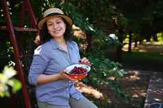 美丽的高加索人女人坐着梯吃甜蜜的成熟的樱桃樱桃果园相机