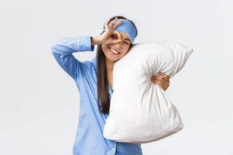 快乐的热情的美丽的亚洲女孩蓝色的睡衣睡觉面具拥<strong>抱枕</strong>头微笑乐观的床上准备好了在外过夜显示手势批准<strong>白色</strong>背景