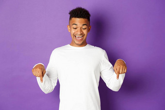 肖像兴奋快乐非裔美国人男人。白色运动衫指出手指微笑惊讶太棒了促销提供站紫色的背景