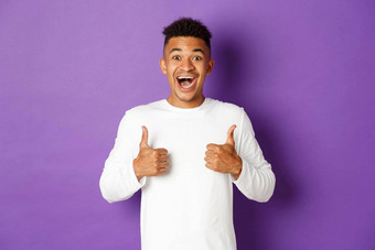 兴奋非裔美国人男人。显示竖起大拇指微笑惊讶赞美伟大的促销站紫色的背景